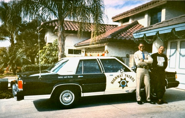 Michael Champion in uniform and his father standing beside a black-and-white CHP Ford LTD Crown Victoria outside a house decorated for Christmas.