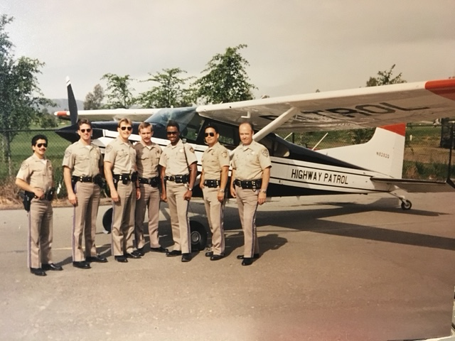 Seven CHP officers in uniform lined up in front of a Highway Patrol Cessna 185 aircraft.
